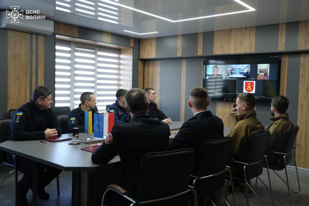 Grupa strażaków uczestniczy w spotkaniu w nowoczesnej sali konferencyjnej. Na stole znajdują się flagi Ukrainy i Unii Europejskiej, a na ekranie widoczna jest wideokonferencja z uczestnikami.